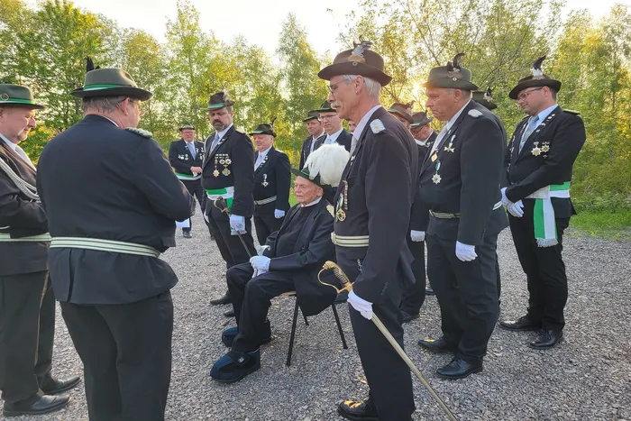 Eine Gruppe von Männern in historischen Uniformen steht um einen älteren Mann, der auf einem Stuhl sitzt. Im Hintergrund Bäume und Strauchwerk.