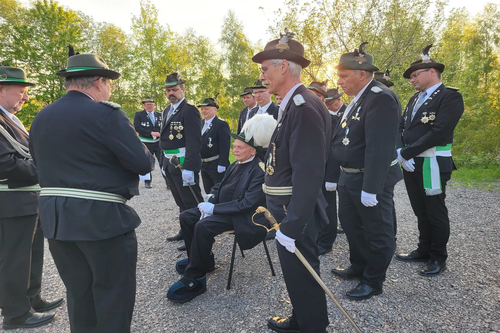 Eine Gruppe von Männern in historischen Uniformen steht um einen älteren Mann, der auf einem Stuhl sitzt. Im Hintergrund Bäume und Strauchwerk.
