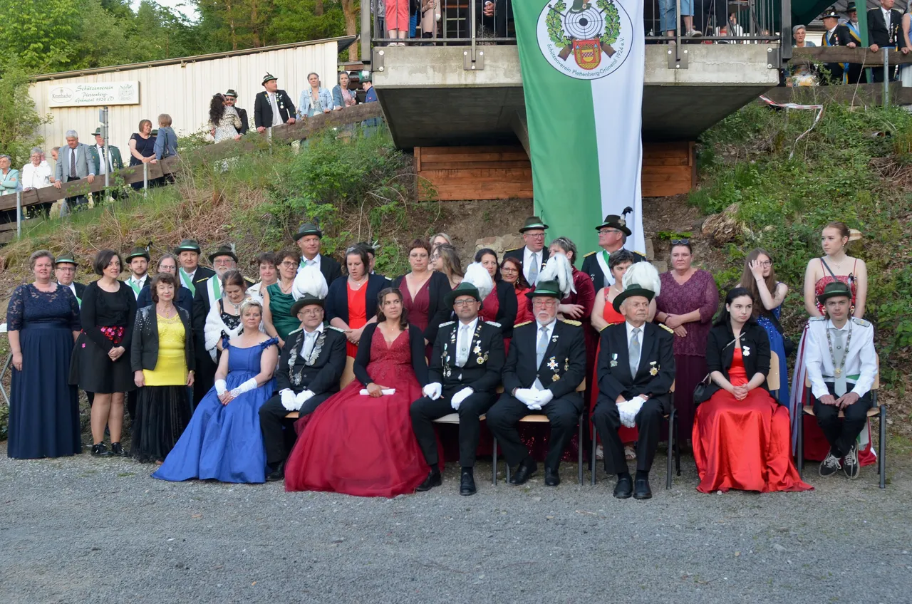 Eine Gruppe von etwa 40 Personen in festlicher Kleidung posiert für ein Foto. Viele tragen traditionelle Schützenuniformen und Orden, einige Frauen sind in langen Abendkleidern zu sehen. Im Hintergrund eine Zuschauermenge und ein Banner mit Wappen.