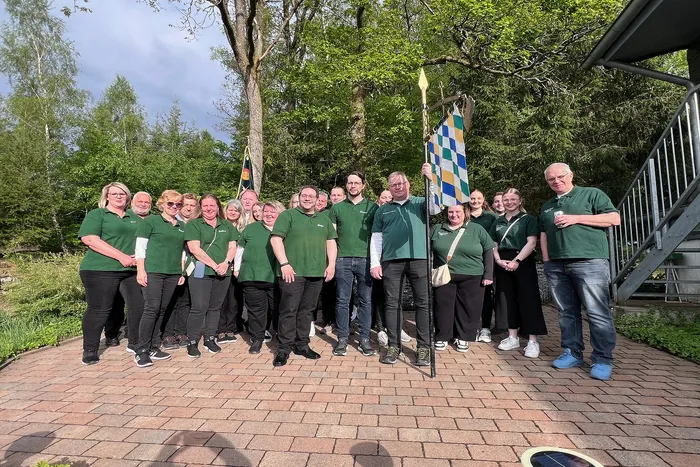 Eine Gruppe von etwa zwanzig Personen in grünen Poloshirts steht auf einem gepflasterten Weg vor Bäumen. Zwei halten Fahnen mit kariertem Muster. Im Hintergrund ein Gebäude mit einem metallenen Geländer.