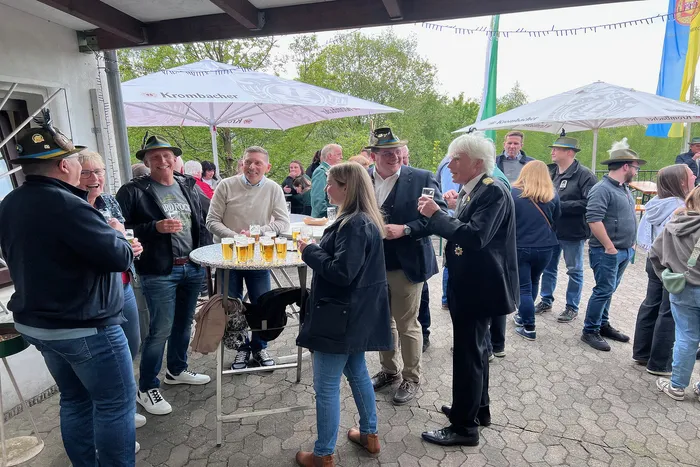 Eine Gruppe von Menschen steht um einen Tisch mit Biergläsern im Freien unter Sonnenschirmen mit Werbeaufdruck. Einige Personen unterhalten sich angeregt, während andere Getränke halten.