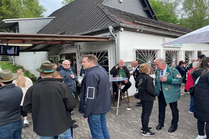 Menschen stehen vor einem Gebäude mit dem Schriftzug „Gasthof Schützenheim“ und unterhalten sich. Im Vordergrund ein Mann mit einem Smartphone in der Hand.