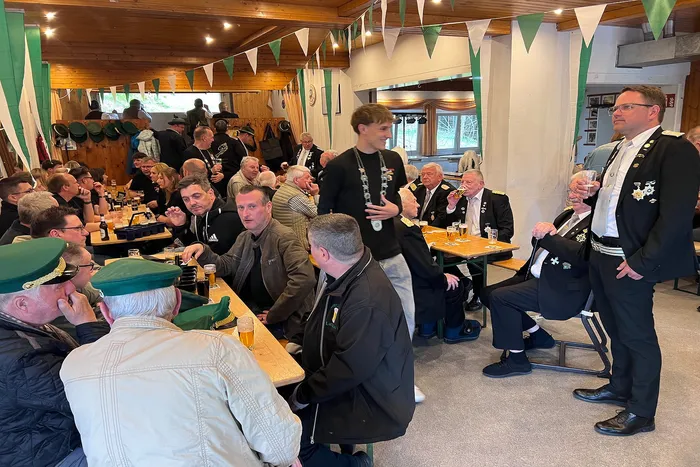 Verschiedene Männer sitzen und stehen in einem hellen Raum mit Holzwänden und Bierkrügen auf Tischen. Im Hintergrund sind grüne Wimpel zu sehen.