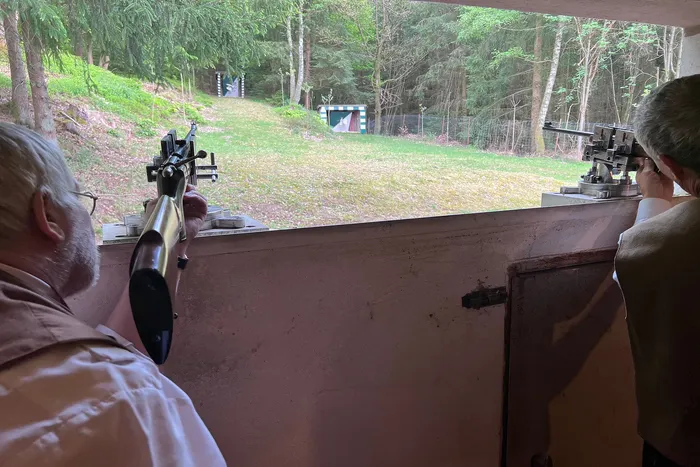 Zwei Männer bedienen aus einem Bunker Gewehre, die auf eine Zielscheibe im Freien gerichtet sind. Im Hintergrund ein Wald.