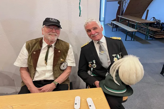 Zwei lächelnde Männer in traditioneller Tracht sitzen an einem Tisch mit Fernbedienungen. Rechts steht ein Hut mit Federn.