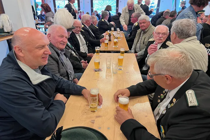 Mehrere ältere Männer sitzen an langen Holztischen in einem hellen Raum und halten Biergläser in der Hand. Im Hintergrund weitere Personen und Tische, die Atmosphäre deutet auf ein geselliges Beisammensein hin.