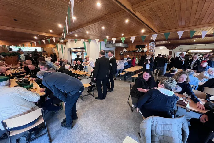 Innenaufnahme eines festlich geschmückten Saals mit vielen Menschen an Tischen, während im Hintergrund uniformierte Schützen stehen. Einige Personen sitzen und unterhalten sich, andere stehen und bewegen sich im Raum.