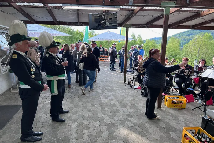 Zwei Männer in historischen Uniformen mit hohen Hüten stehen im Vordergrund, während im Hintergrund Menschen auf einer überdachten Fläche stehen und eine Blaskapelle spielt.