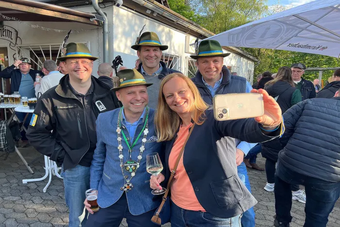 Fünf Männer und eine Frau in Tracht posieren lächelnd für ein Selfie. Die Frau hält ein Weinglas, einer der Männer hält eine Schießscheibe. Im Hintergrund sind weitere Menschen und ein Festzelt zu sehen.