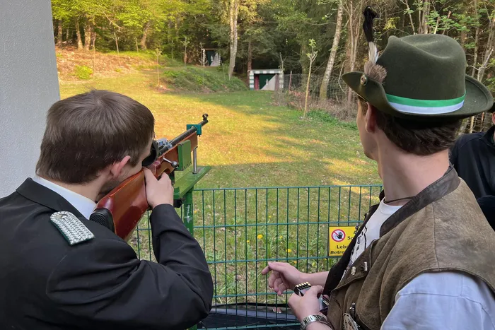Zwei Männer bei einem Schützenfest: Ein Mann in Uniform zielt mit einem Gewehr, während ein anderer Mann mit Hut zusieht. Im Hintergrund ein grüner Zaun und Bäume.