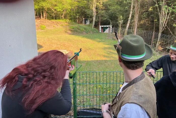 Eine Frau zielt mit einem Gewehr auf eine Zielscheibe, während ein Mann mit Hut und ein weiterer Beobachter zusehen. Im Hintergrund eine grüne Landschaft und ein Schützenstand.