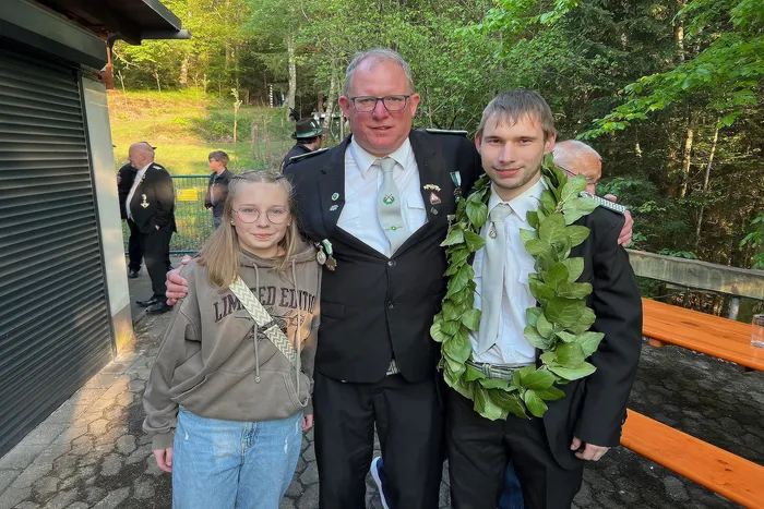 Drei Personen posieren für ein Foto: ein Mann in Schützenuniform, ein Mädchen und ein junger Mann mit einem grünen Kranz um den Hals. Im Hintergrund sind weitere Schützen und ein Wald zu sehen.