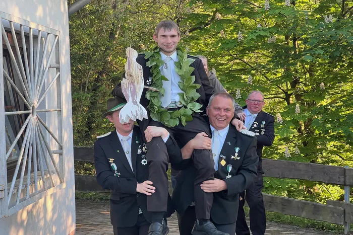 Drei Männer tragen einen jüngeren Mann auf ihren Schultern, der mit einem Kranz geschmückt ist. Alle tragen Schützenuniformen mit Orden und Abzeichen.