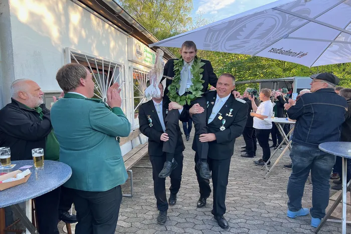 Vier Männer tragen einen jüngeren Mann auf ihren Schultern, während dieser einen grünen Kranz hält. Im Hintergrund steht ein Gebäude mit einem Schild und Menschen stehen um Tische.