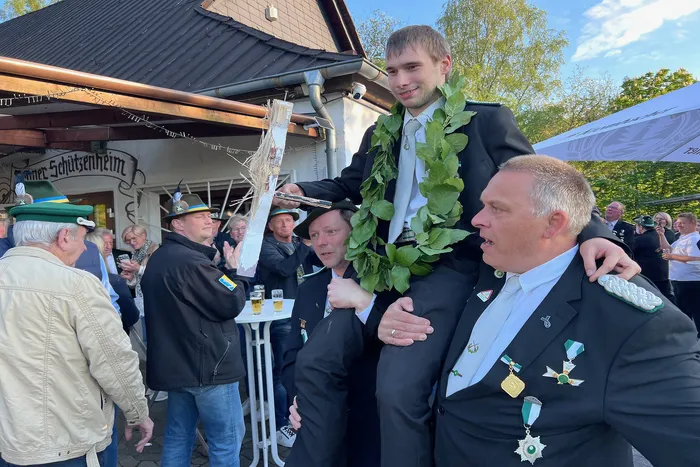 Junge erwachsene Person mit Blumenkranz um den Hals wird von zwei älteren Männern in Anzug und mit Orden begleitet, im Hintergrund ein Schützenverein mit dem Schriftzug „Erner Schützenheim“.