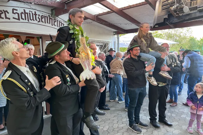 Junge erwachsene Person wird auf den Schultern eines Mannes getragen, während ein Mann in Uniform und ein anderer mit Kind zusehen. Im Hintergrund ein Gebäude mit der Aufschrift „Schützenheim“.