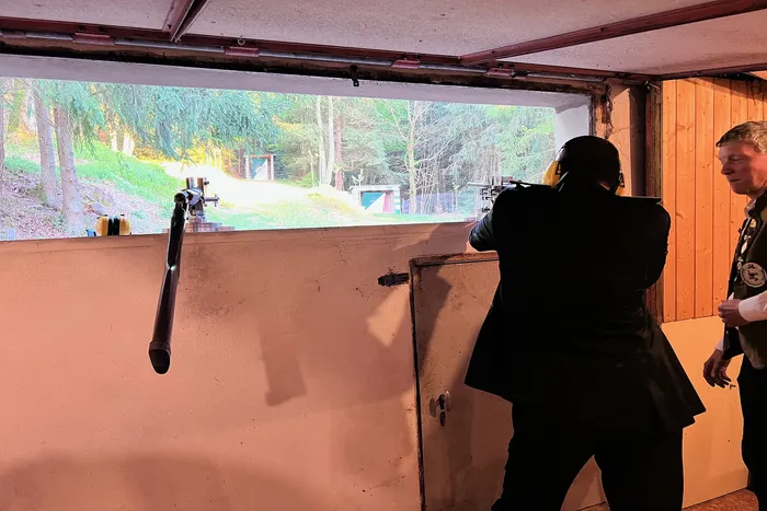 Ein Schütze zielt mit einer Waffe auf eine Zielscheibe in einem Schießstand, während ein weiterer Mann ihm zusieht. Im Hintergrund ist eine bewaldete Landschaft durch eine große Öffnung sichtbar.