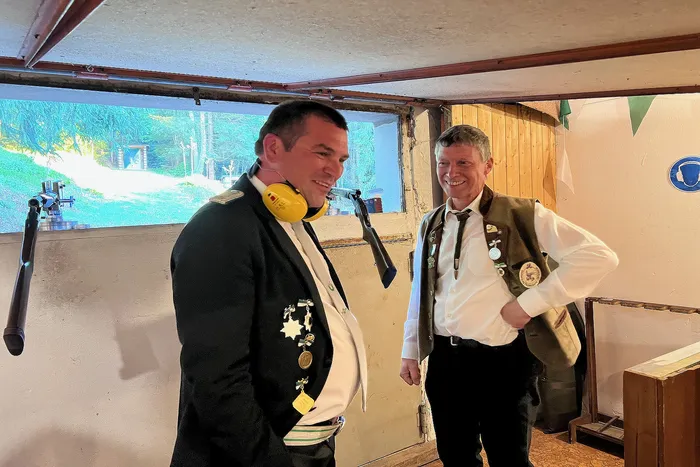 Zwei Männer in traditioneller Schützenkleidung lächeln in einem Raum mit Fenstern zu einer grünen Landschaft. Einer trägt Gehörschutz.