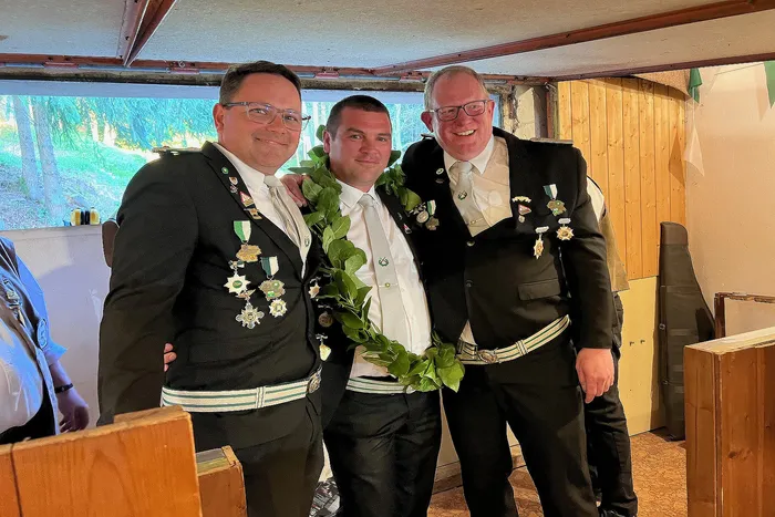 Drei Männer in traditioneller Schützenkleidung, darunter der frischgekrönte Schützenkönig Christian Hoffmann mit einem grünen Kranz um den Hals, posieren lächelnd für ein Foto.
