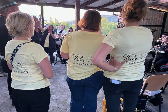 Drei Personen mit gelben T-Shirts, auf denen „Tada, da sind wir wieder“ steht, stehen dem Betrachter den Rücken zu. Im Hintergrund spielt eine Blaskapelle unter einem hölzernen Pavillon.