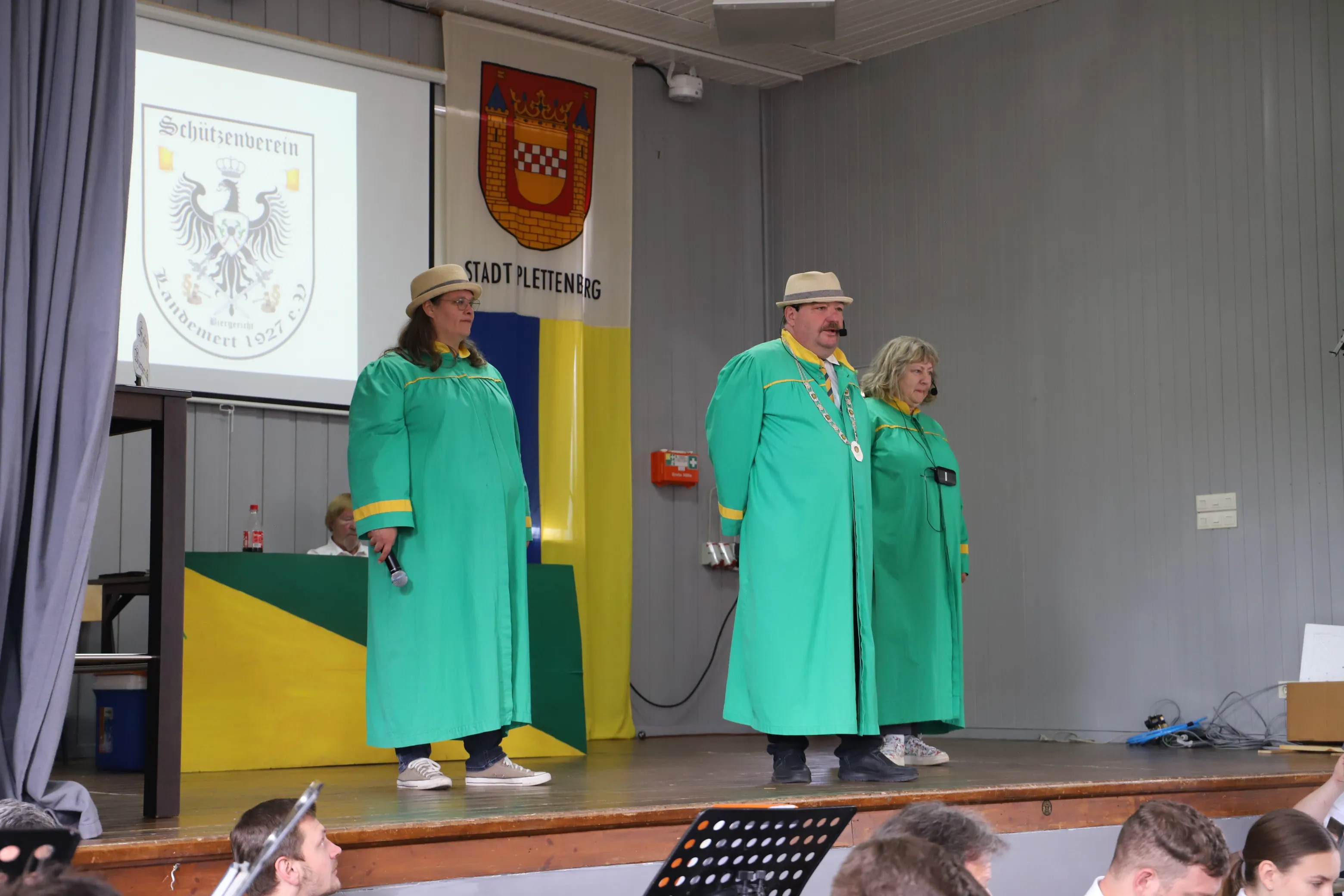 Zwei Personen in grünen Gewändern und eine dritte in ähnlicher Kleidung mit Zylinder stehen auf einer Bühne vor einem Publikum. Im Hintergrund sind Wappen und ein Adler zu sehen.