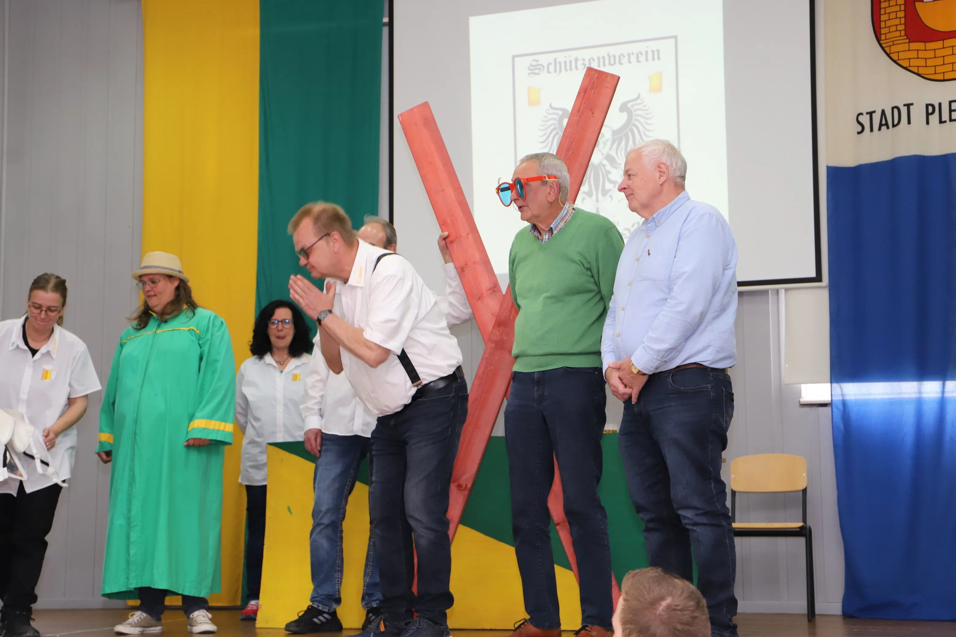Drei Männer werden auf einer Bühne von zwei weiteren Personen mit Holzbalken gekreuzigt, während sie sich an den Kopf fassen oder eine Schutzbrille tragen. Im Hintergrund sind Banner und das Stadtwappen zu sehen.