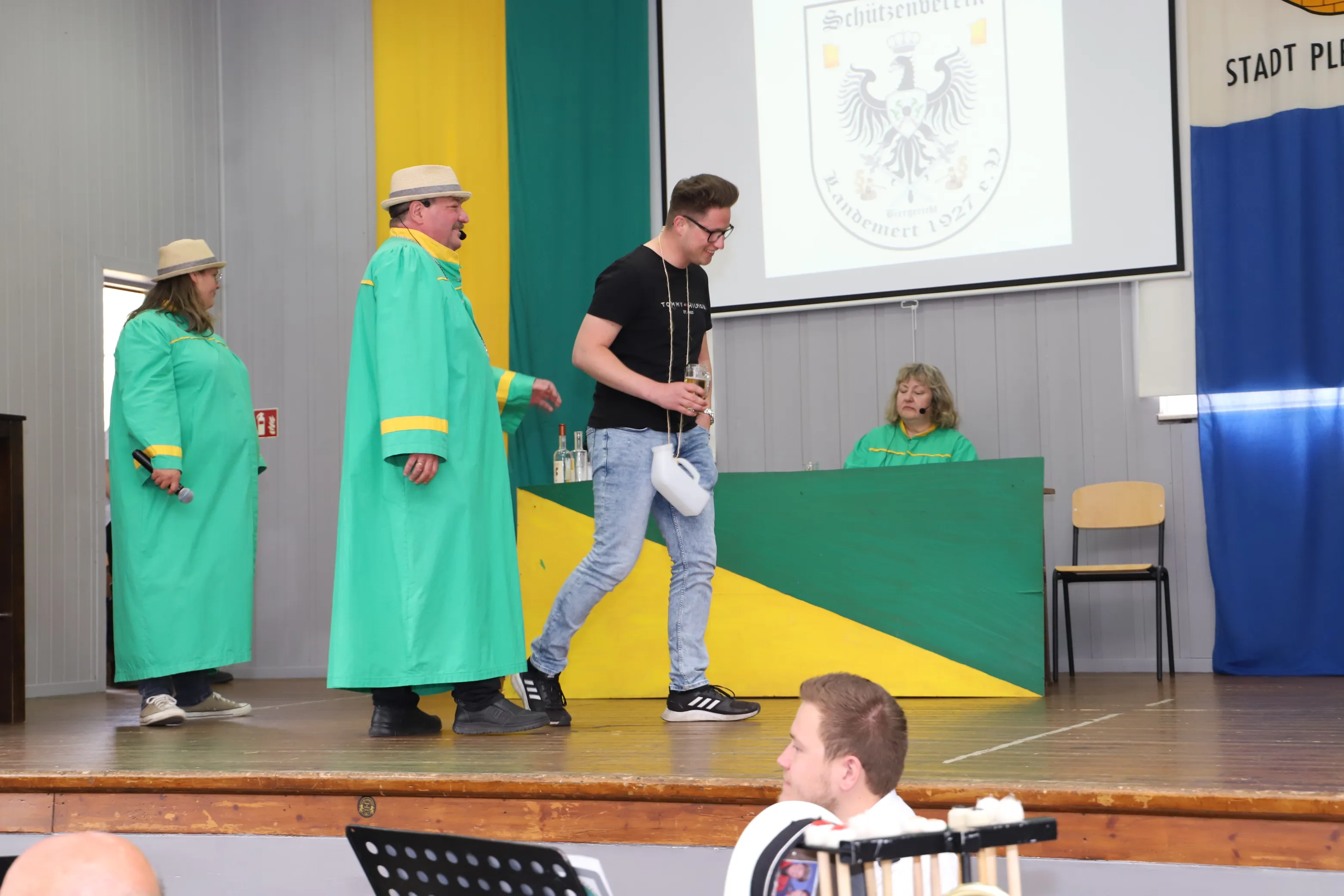 Drei Schützen in historischen Gewändern präsentieren einen jüngeren Mann auf einer Bühne, während im Hintergrund ein Wappen projiziert wird. Das Publikum verfolgt das Geschehen von ihren Plätzen aus.
