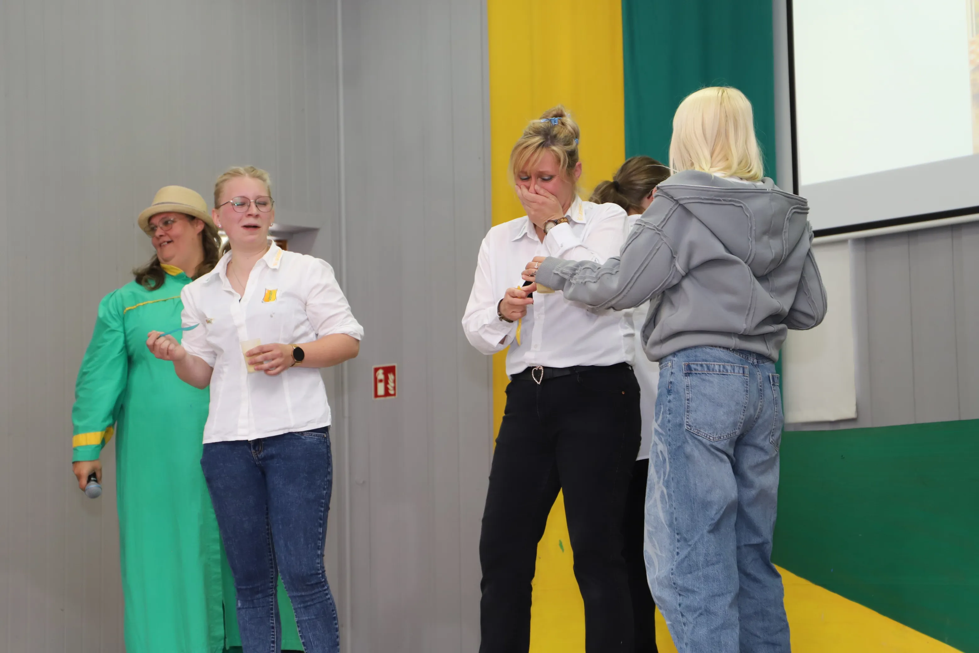 Drei Frauen stehen auf einer Bühne, während eine vierte im Hintergrund steht. Die Frau links trägt ein grünes Kleid, die mittlere eine weiße Bluse und Jeans, und die rechts weint und wird von einer weiteren Frau getröstet.