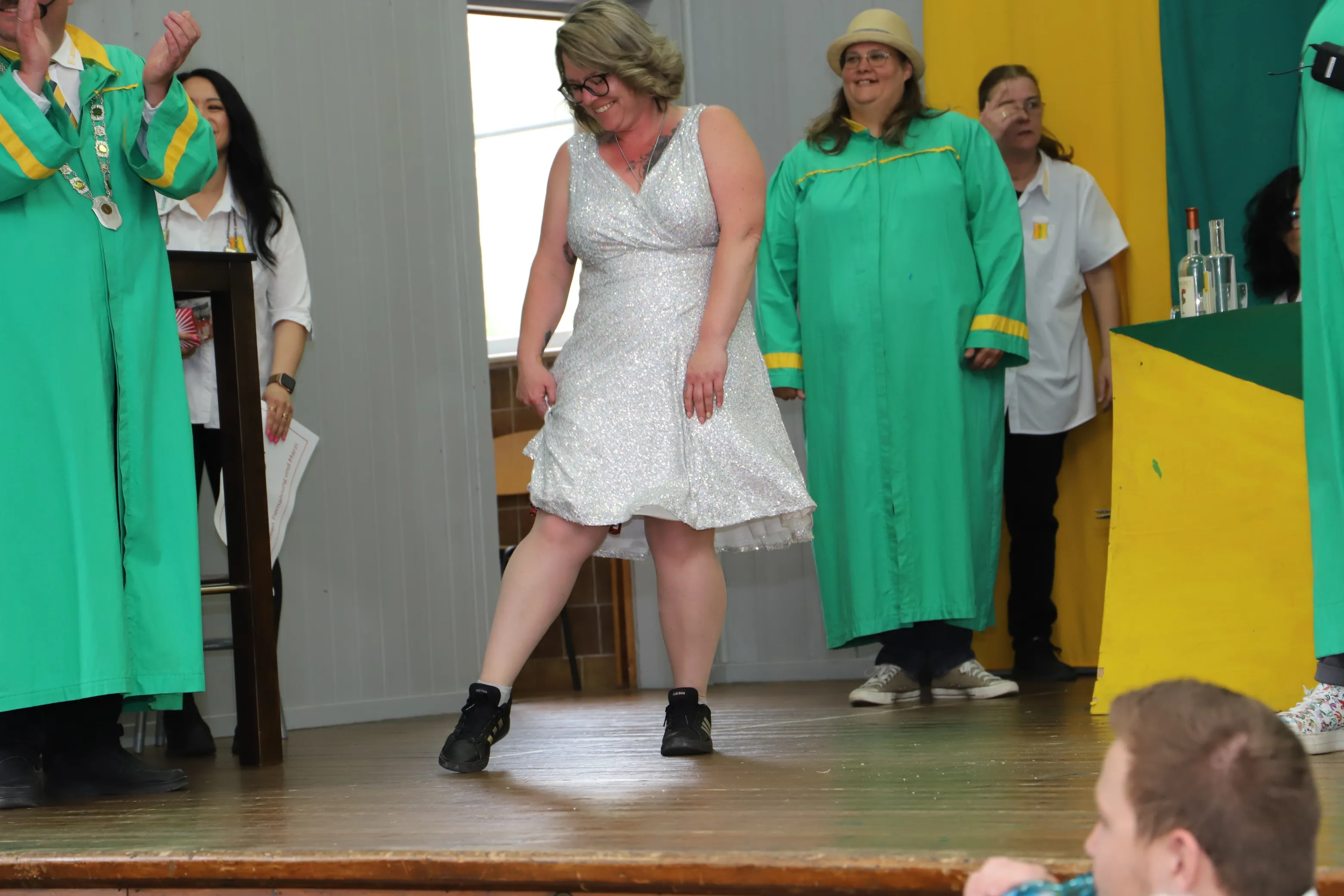 Eine Frau in silbernem Kleid und Turnschuhen steht auf einer Bühne und blickt lächelnd ins Publikum. Um sie herum stehen Personen in grünen Umhängen.