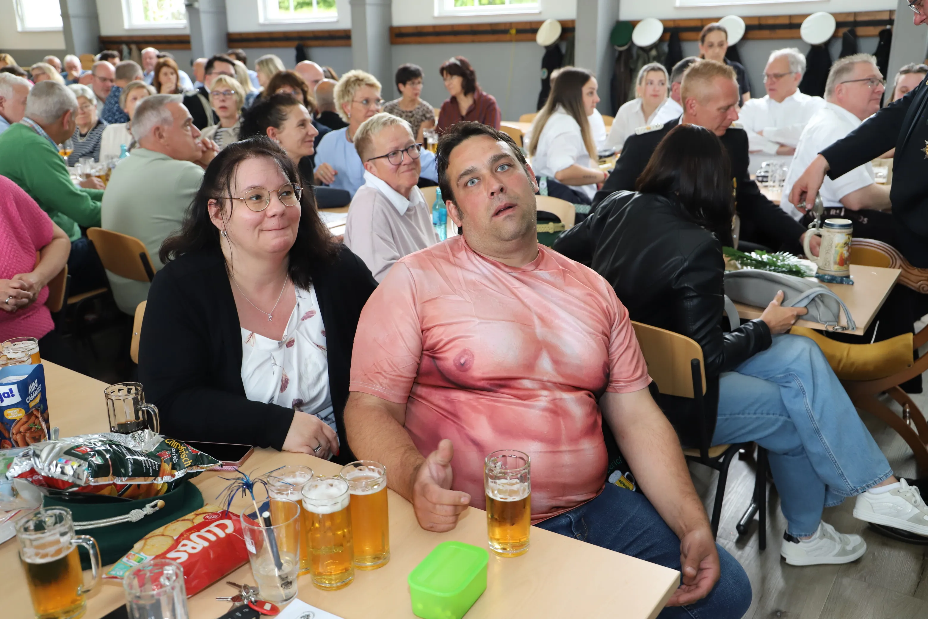 Zwei Zuschauer, eine Frau und ein Mann, blicken aufmerksam nach vorn, während um sie herum viele Menschen an Tischen sitzen und Bier trinken. Im Hintergrund eine Bühne.
