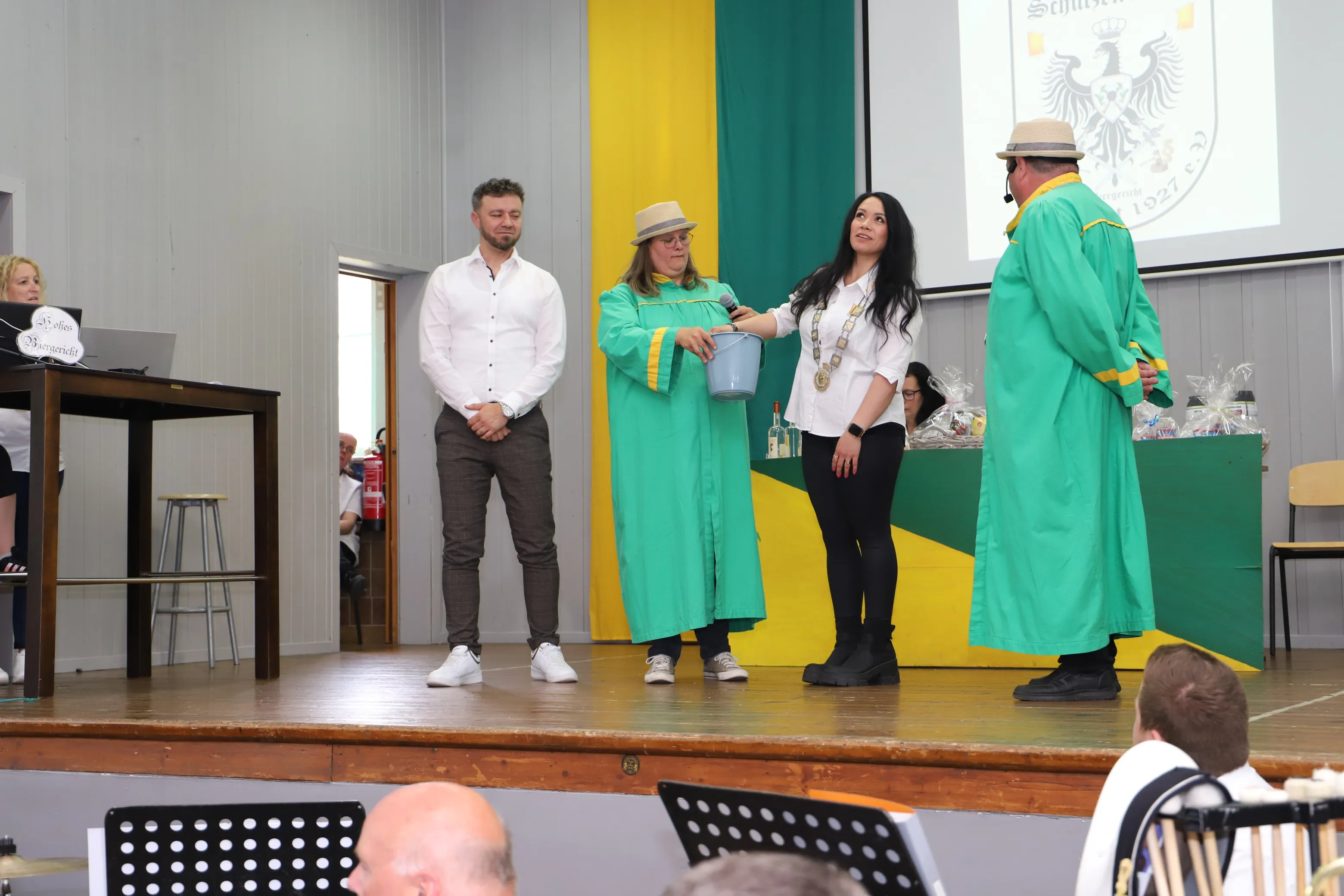 Zwei Männer und eine Frau in traditionellen Schützenumhängen stehen auf einer Bühne vor einem Publikum. Die Frau hält einen Eimer und eine Kette um den Hals.
