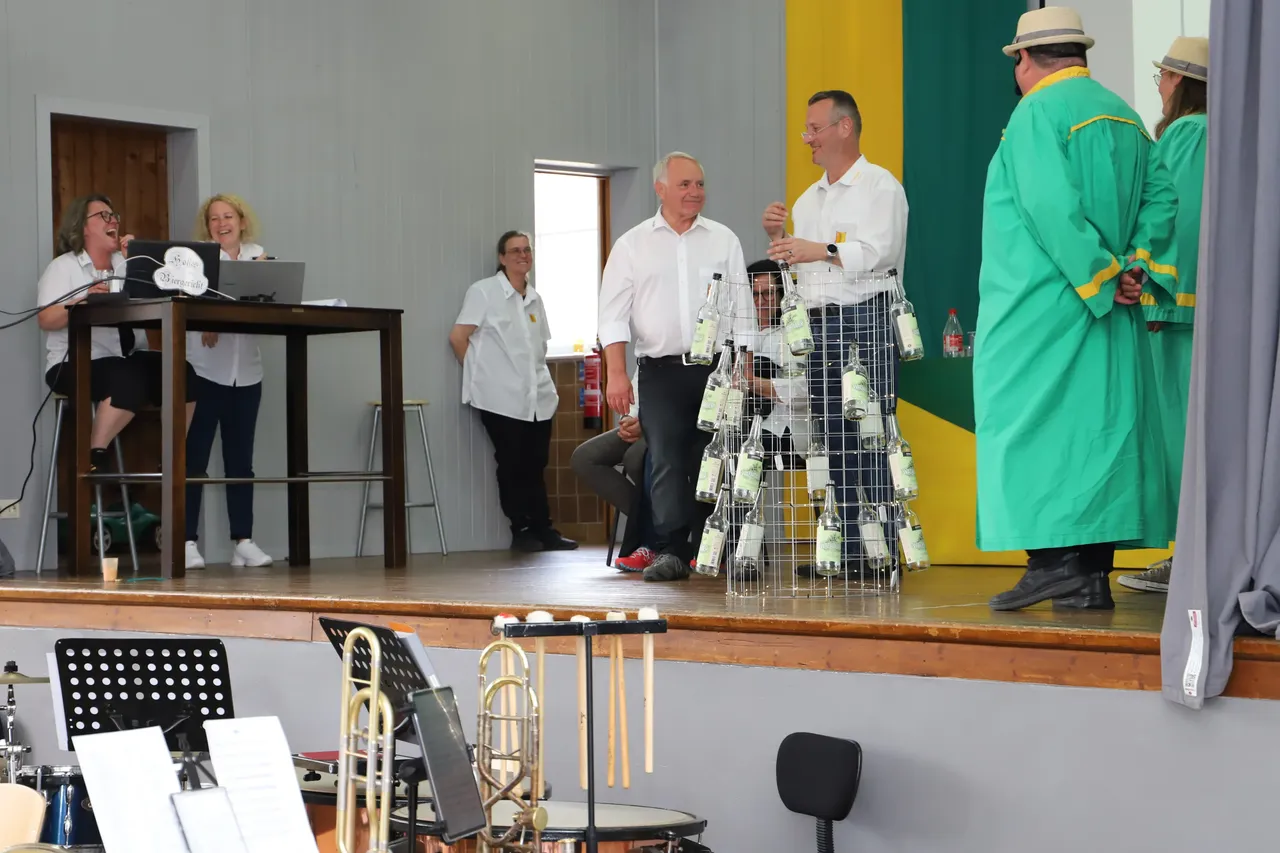 Auf einer Bühne stehen mehrere Männer in weißen Hemden und ein Mann in grüner Robe, während eine Frau im Hintergrund am Keyboard spielt. Vor der Bühne sind Musikinstrumente wie Posaunen und ein Schlagzeug zu sehen.
