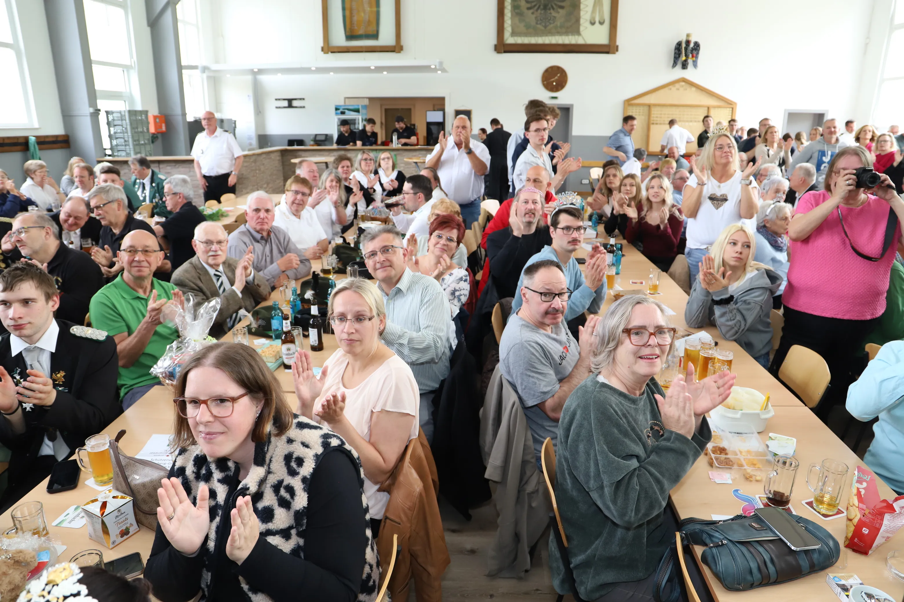 Große Menschenmenge in einem hellen Saal klatscht und lacht, während einige Personen auf einer kleinen Bühne stehen. Biere und Snacks stehen auf den Tischen im Vordergrund.