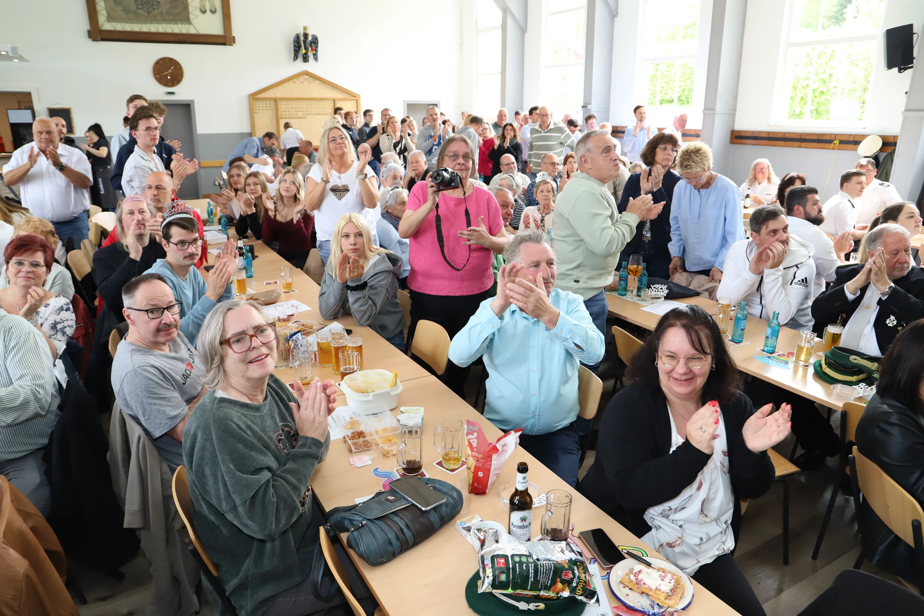 Zuschauer klatschen und lächeln in einem hellen Saal mit langen Biertischen. Auf den Tischen stehen Getränke und Snacks.