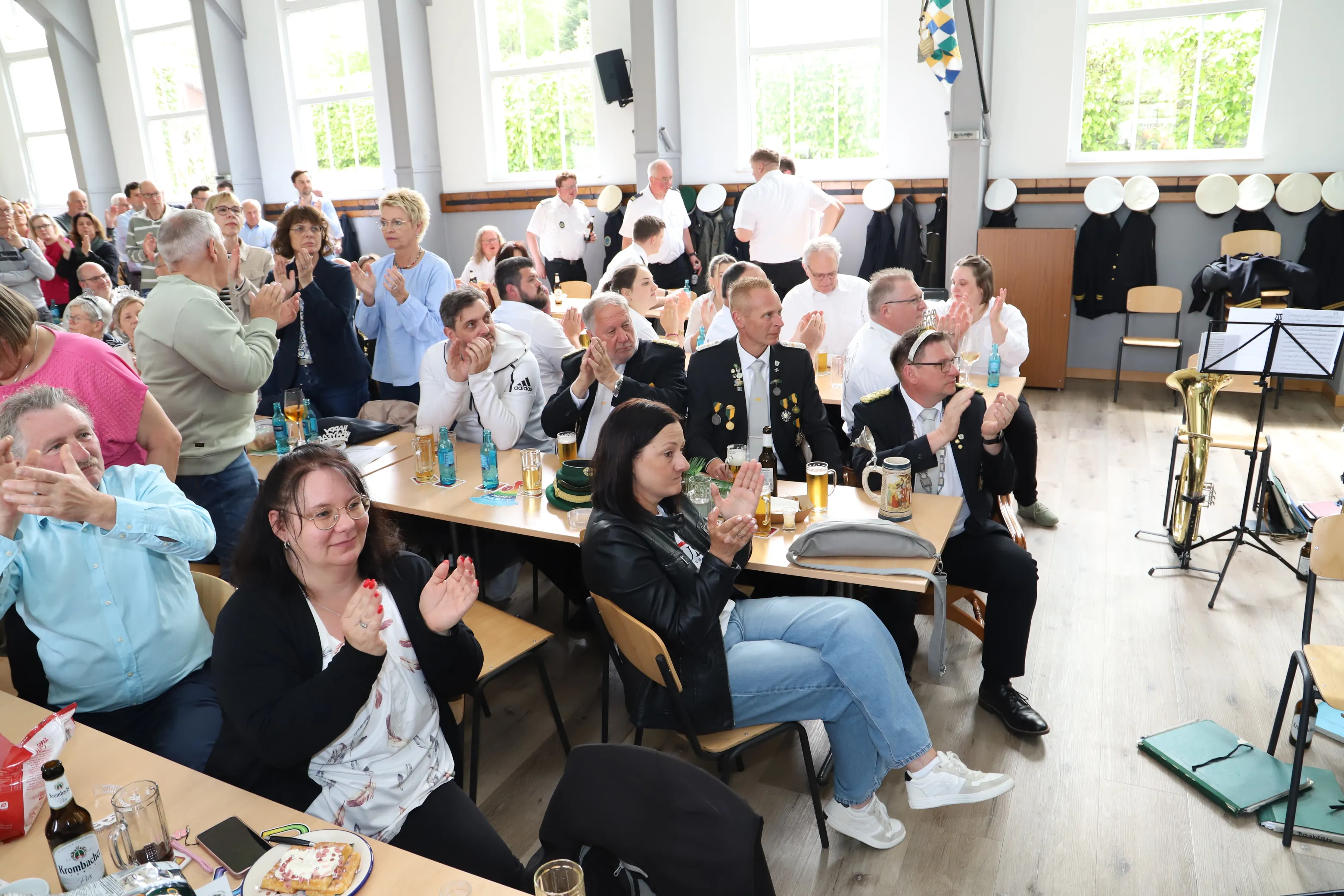 Zuschauer klatschen und lachen während einer Veranstaltung in einem hellen Saal mit langen Tischen und Bierkrügen.