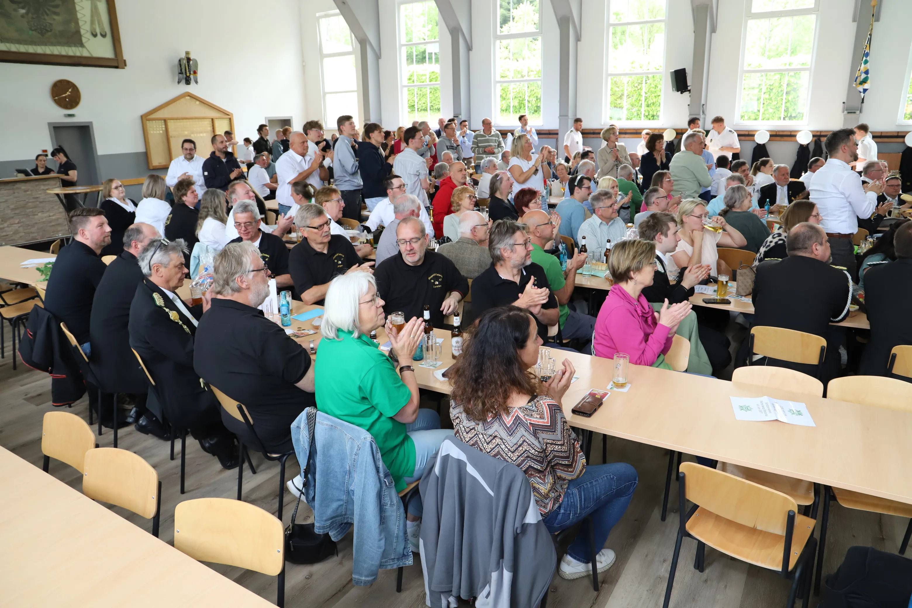 Große Menschenmenge in einem hellen Saal, viele sitzen an Tischen mit Bierkrügen und verfolgen eine Präsentation auf einer kleinen Bühne.