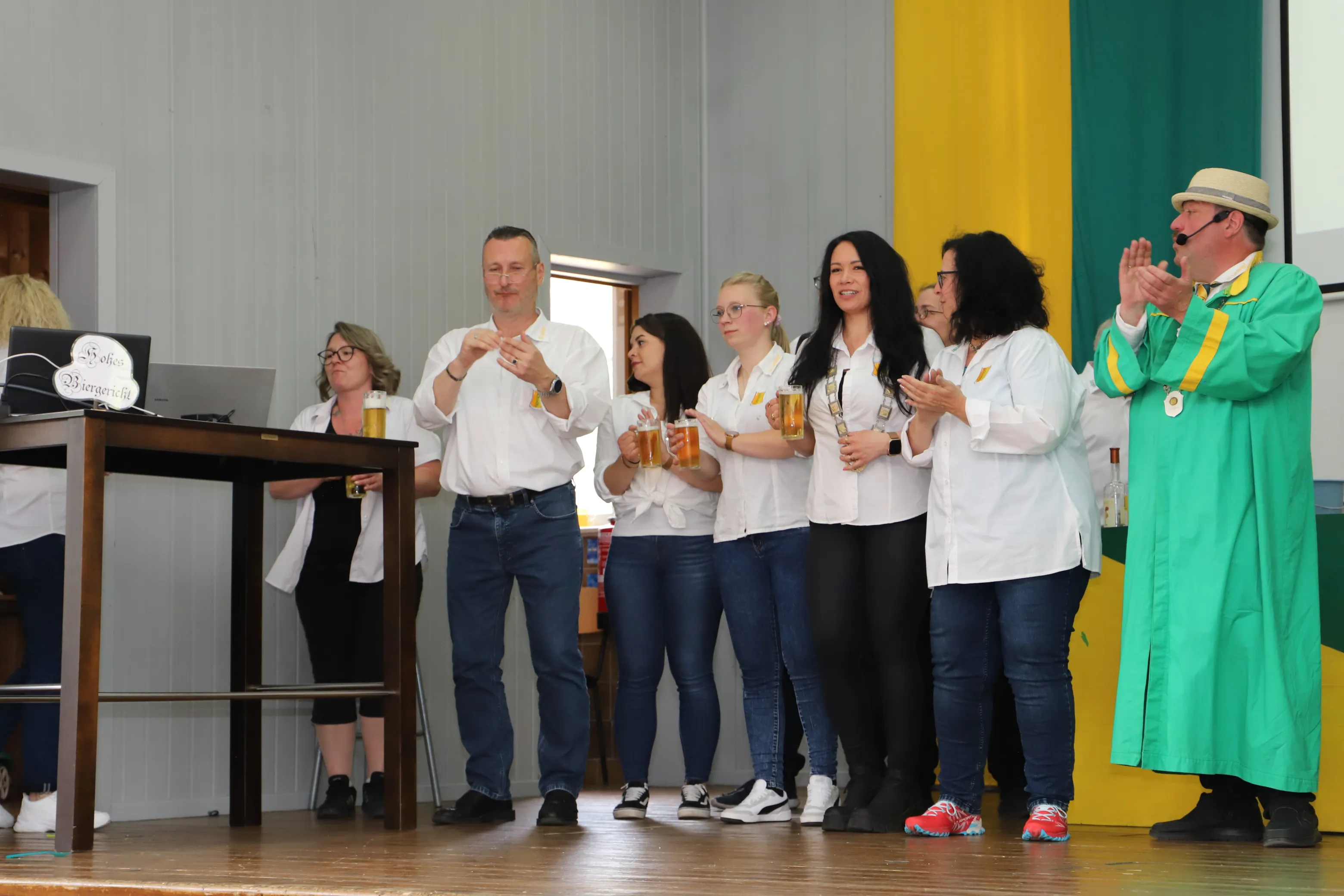 Mehrere Personen auf einer Bühne halten Bierkrüge hoch und klatschen, während ein Mann in grünem Gewand und weißem Hut begeistert applaudiert.