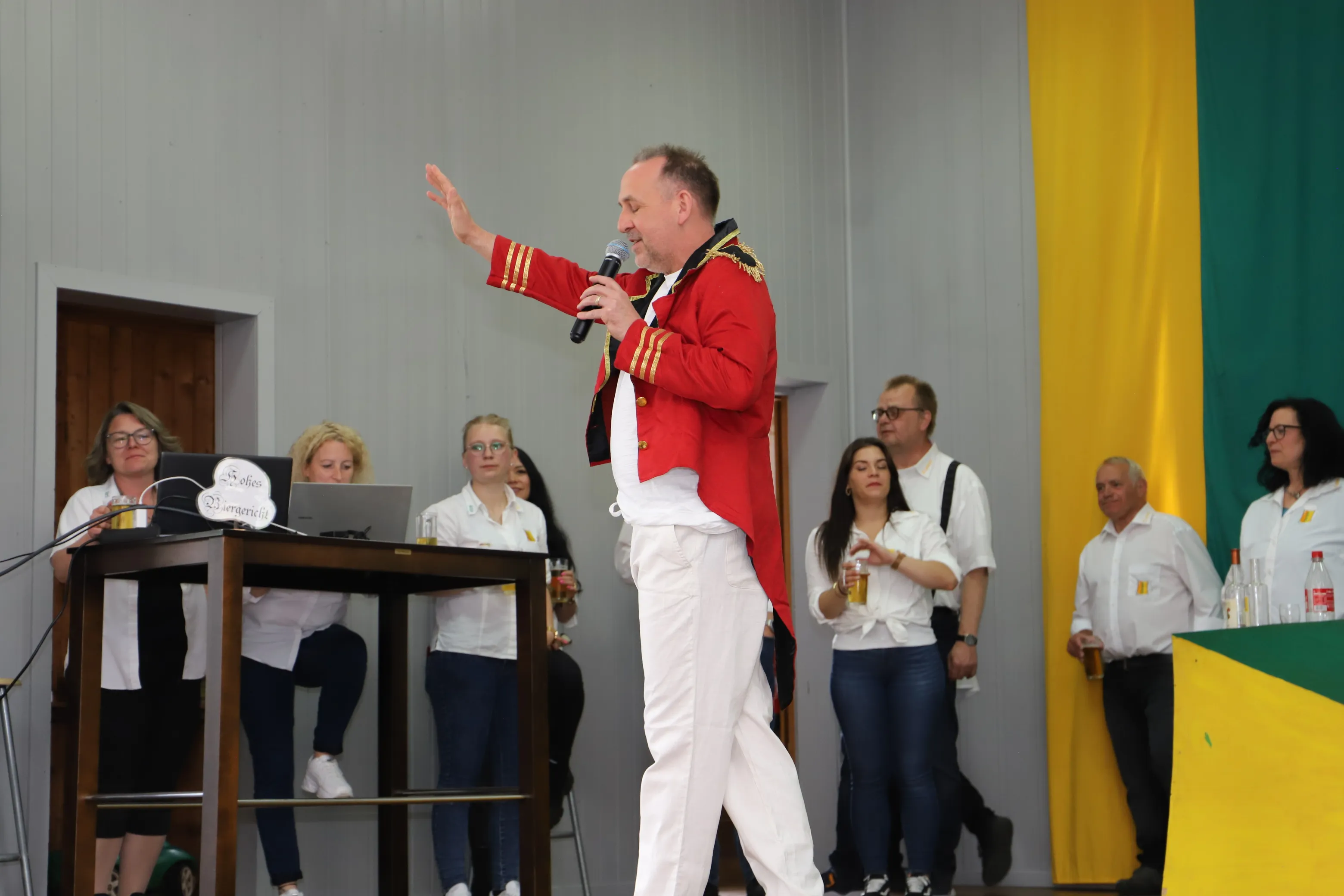 Ein Mann in roter Uniform hält ein Mikrofon und gestikuliert auf einer Bühne, während mehrere Personen im Hintergrund zusehen. Links steht ein Pult mit einem Laptop.