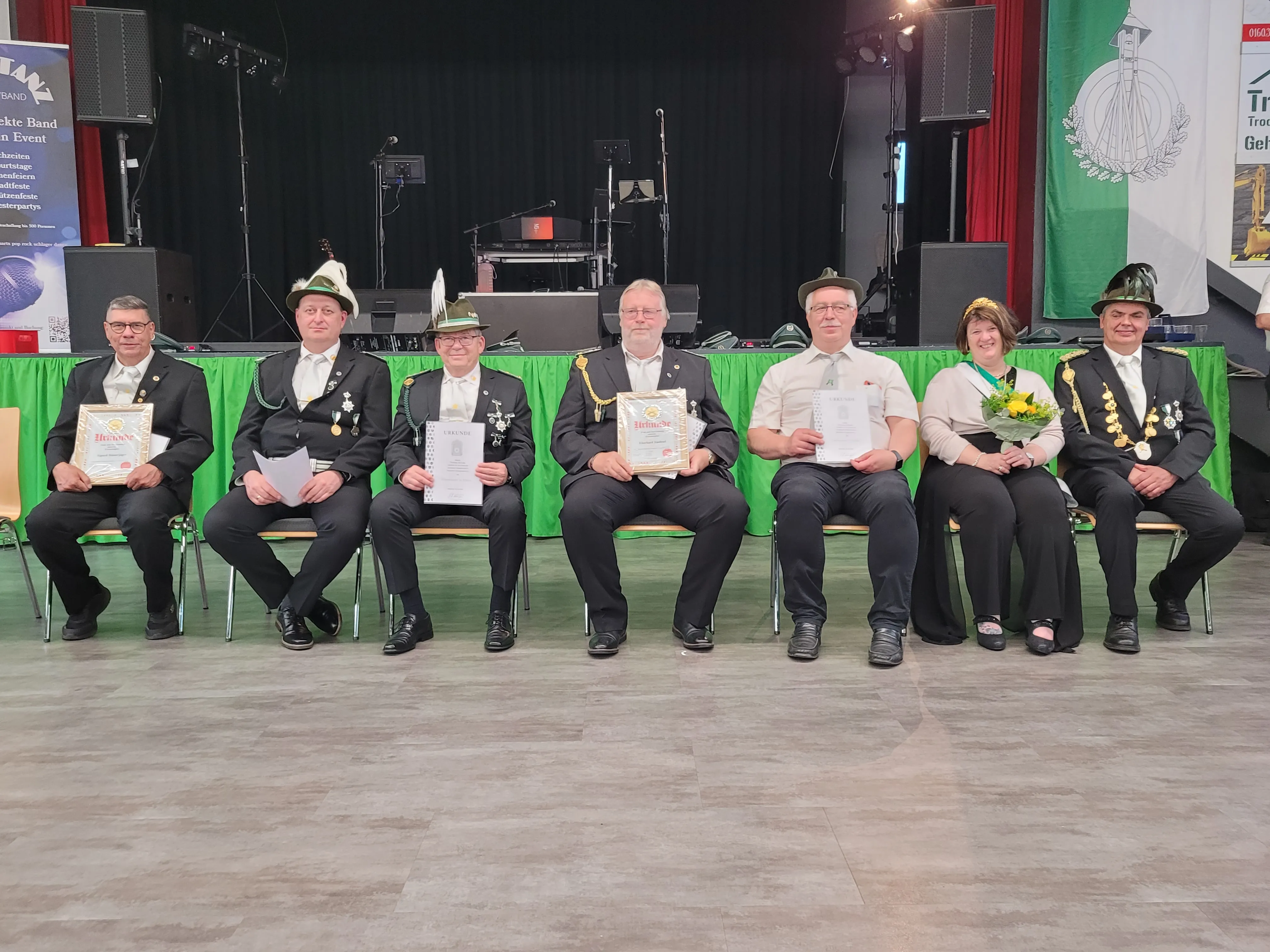 Sieben Personen in traditionellen Schützenuniformen sitzen auf einer Bühne und halten Urkunden. Im Hintergrund eine Bühne und Banner.