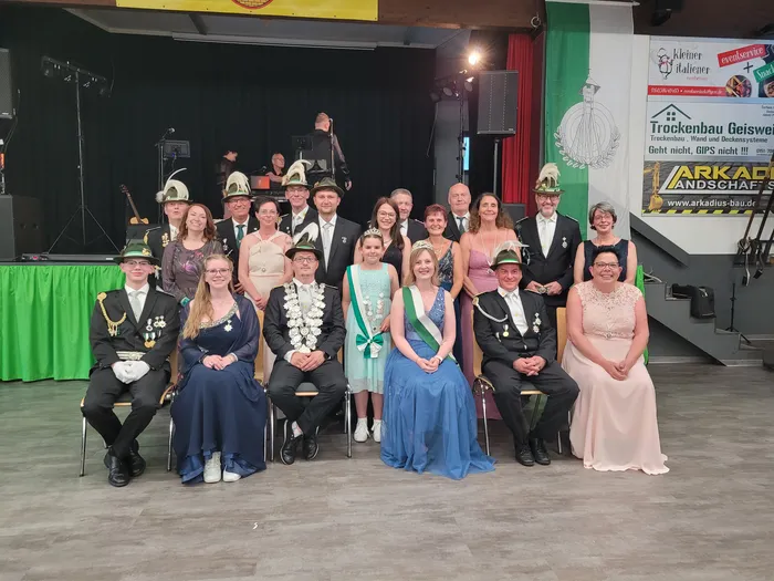 Gruppenfoto von etwa zwanzig Personen in festlicher Kleidung vor einer Bühne mit Werbebanner. Im Mittelpunkt ein Königspaar mit Krone und Ordensband.