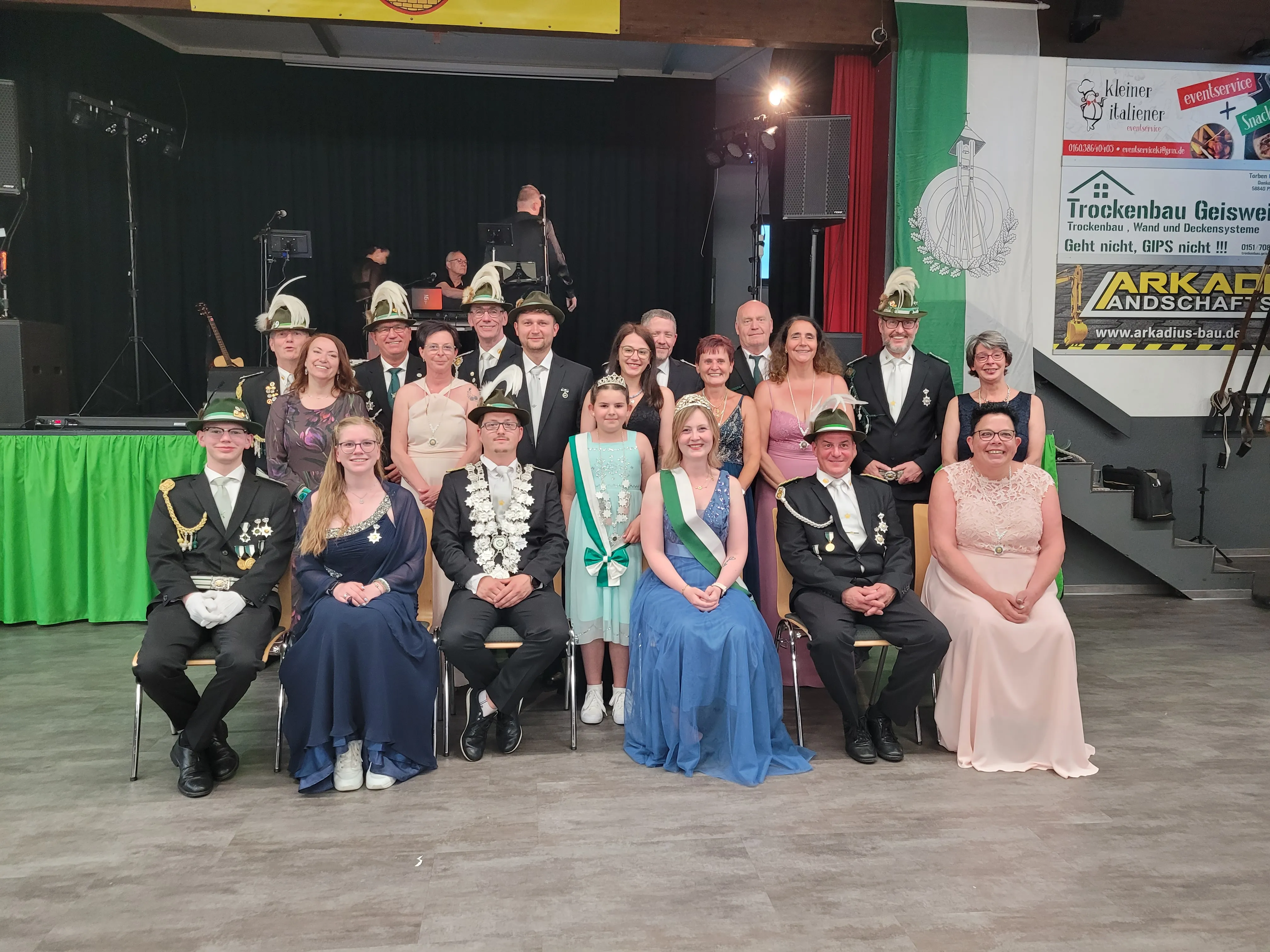 Gruppenfoto von etwa zwanzig Personen in festlicher Kleidung vor einer Bühne mit Werbebanner. Im Mittelpunkt ein Königspaar mit Krone und Ordensband.