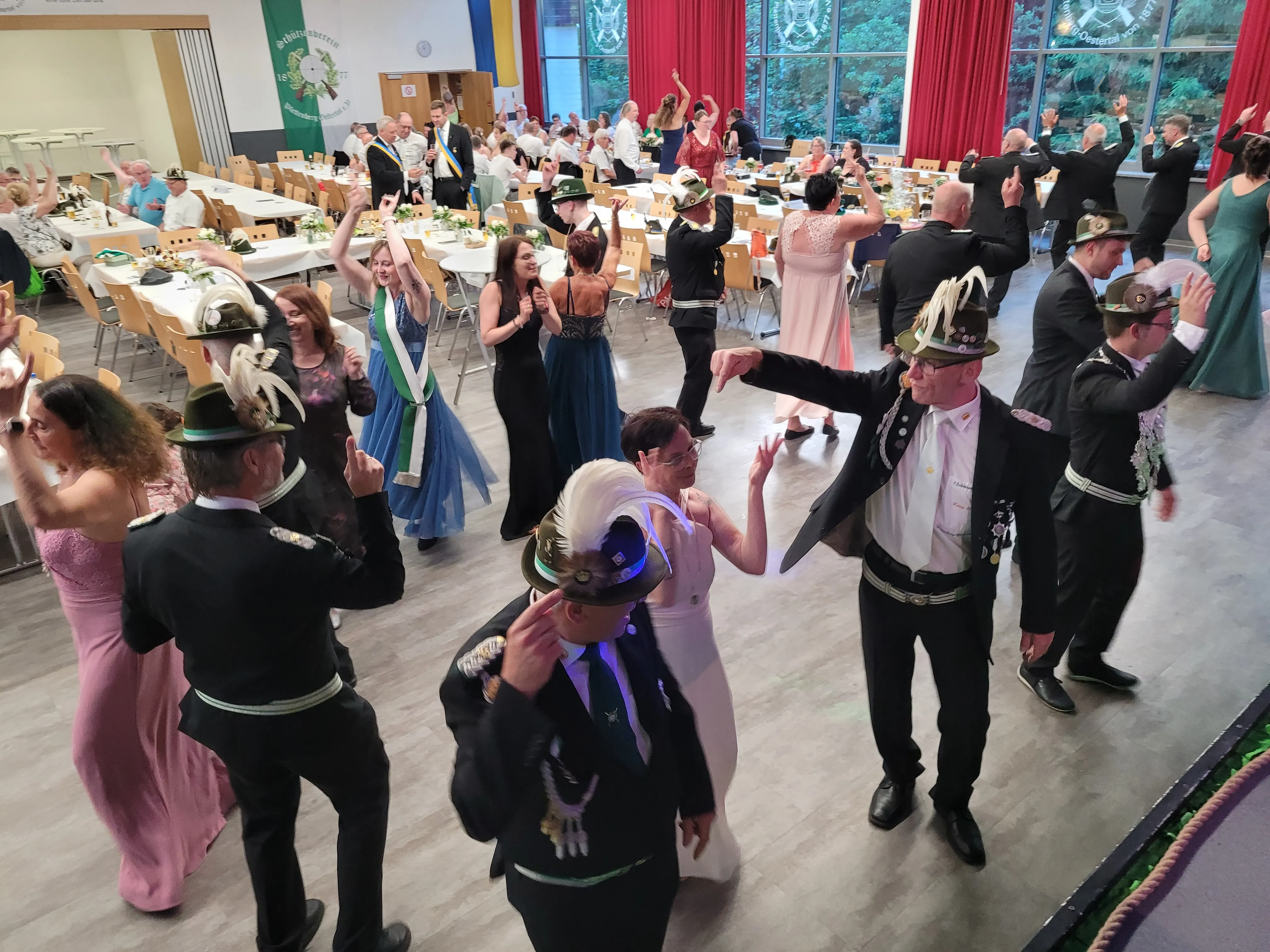 Tanzende Menschen in traditioneller Tracht feiern in einem festlich dekorierten Saal. Im Hintergrund sind gedeckte Tische und Wappen zu sehen.