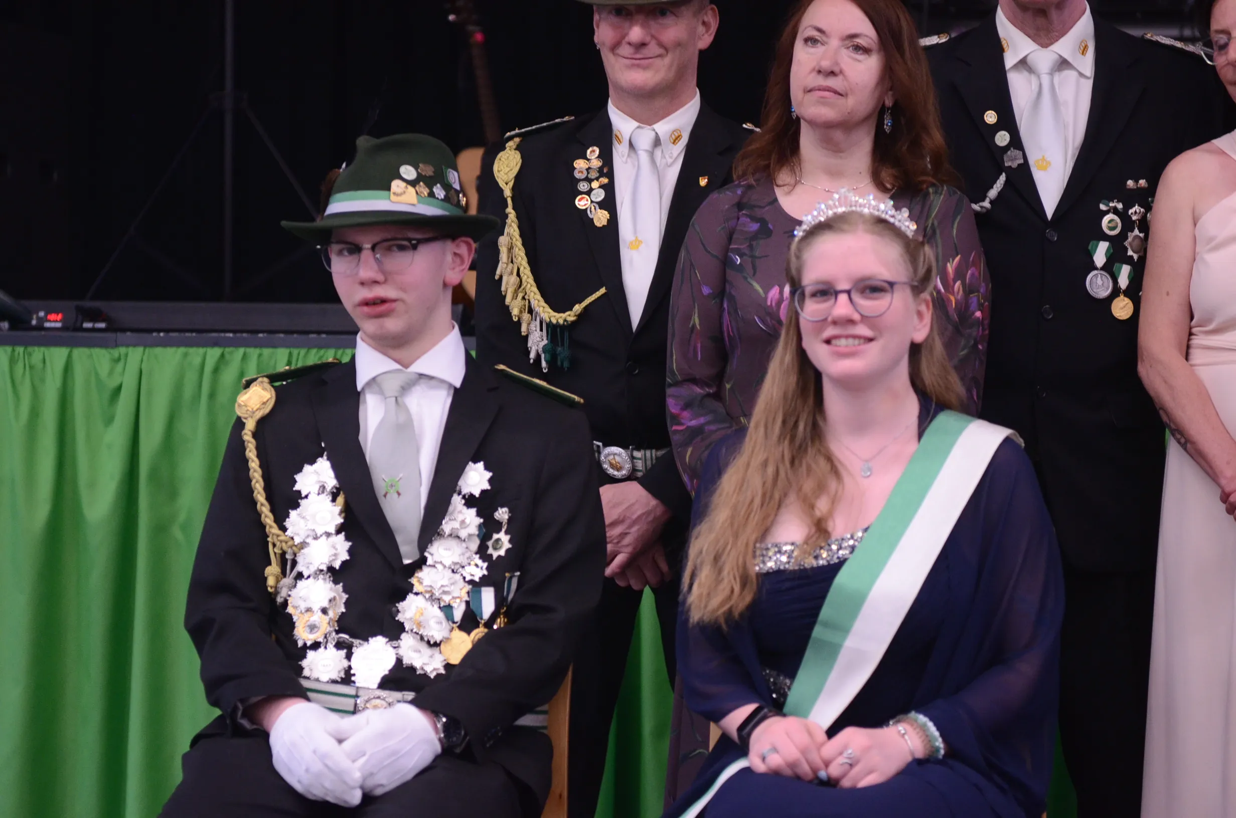 Junge und Mädchen in festlicher Tracht sitzen vor einer Gruppe von Erwachsenen mit Orden und Auszeichnungen. Im Hintergrund eine grüne Bühne.