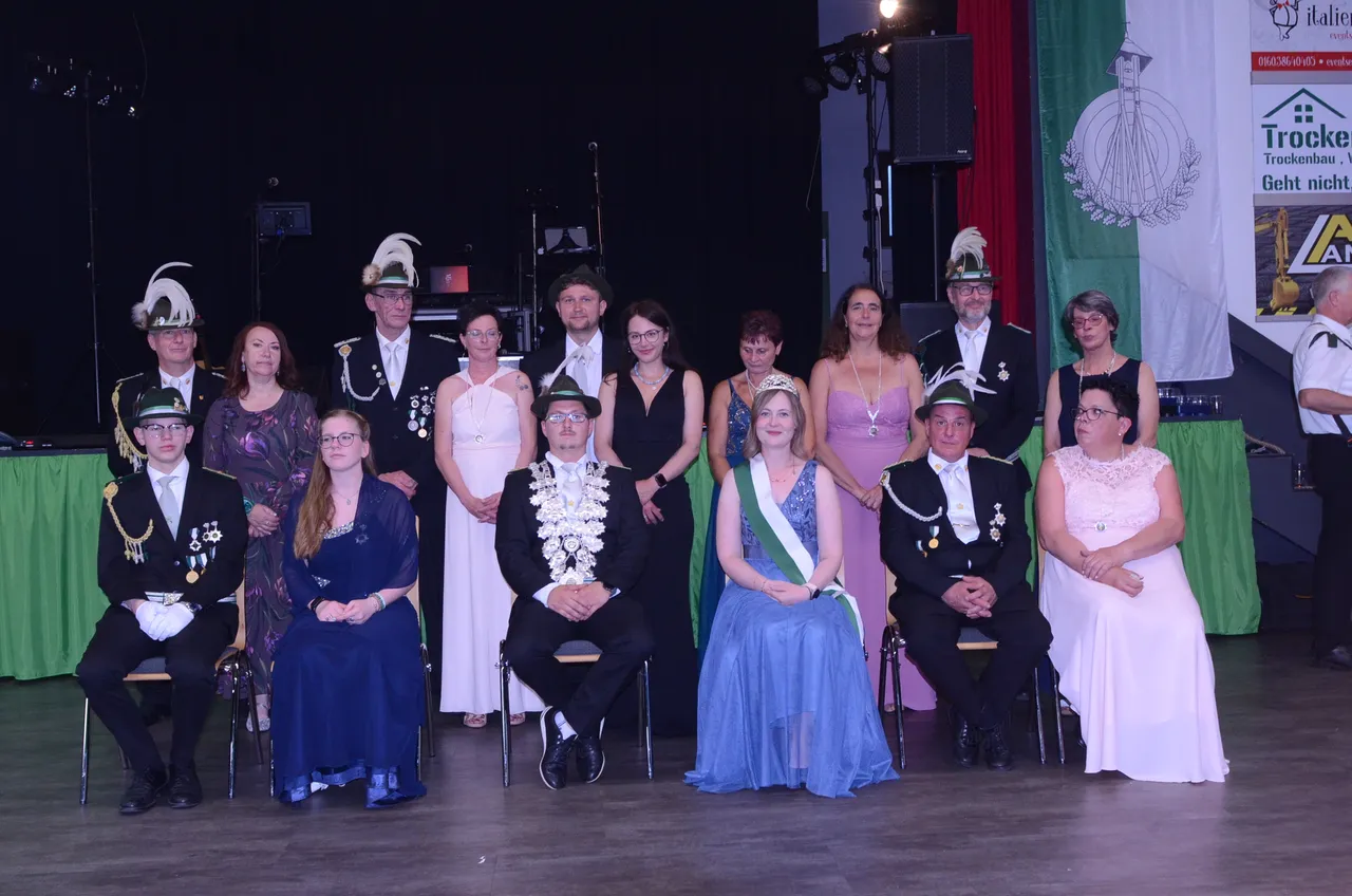 Gruppenfoto von zehn Personen in festlicher Kleidung auf einer Bühne, darunter Mitglieder einer Schützenkompanie und das frisch gekrönte Königspaar mit Krone und Ordensband.