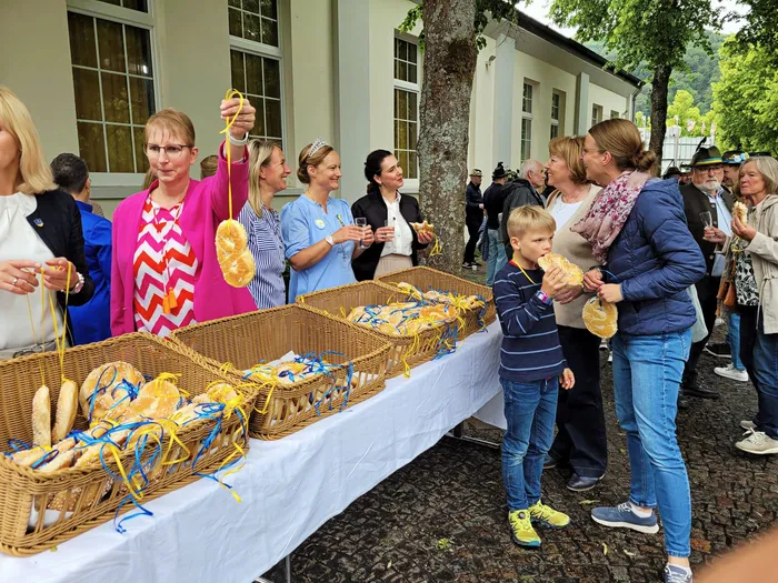 Menschen stehen um einen Tisch mit geflochtenen Körben voller Osterbrot. Ein Junge hält ein Brot hoch, während eine Frau ihm dabei zusieht. Im Hintergrund sind weitere Personen und ein Gebäude zu sehen.