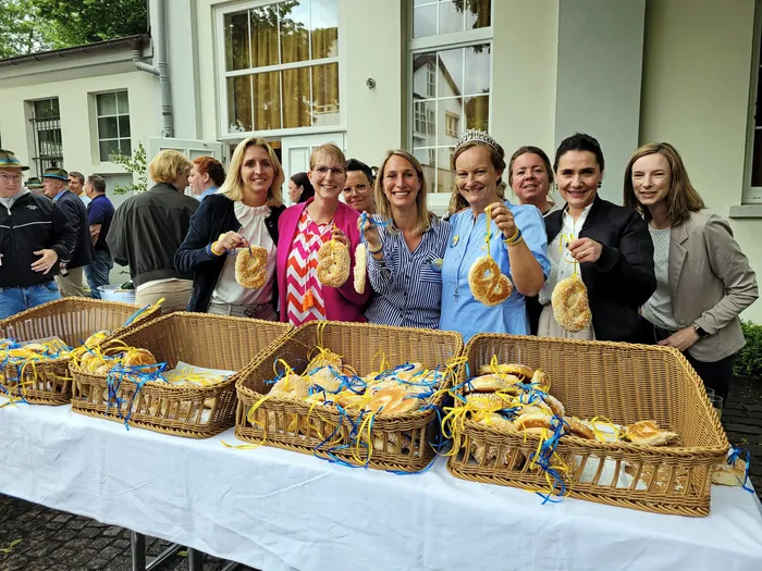 Eine Gruppe von Frauen hält Brezeln vor einem Gebäude mit Zuschauern im Hintergrund. Im Vordergrund stehen Körbe mit Brezeln auf einem Tisch.
