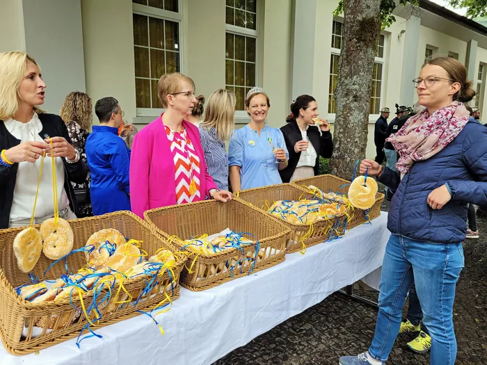 Eine Gruppe von Menschen steht vor einem Gebäude und unterhält sich. Im Vordergrund steht ein Tisch mit Körben voller Osterbrote, von denen einige an Bändern hängen. Eine Frau hält ein Brot in der Hand.