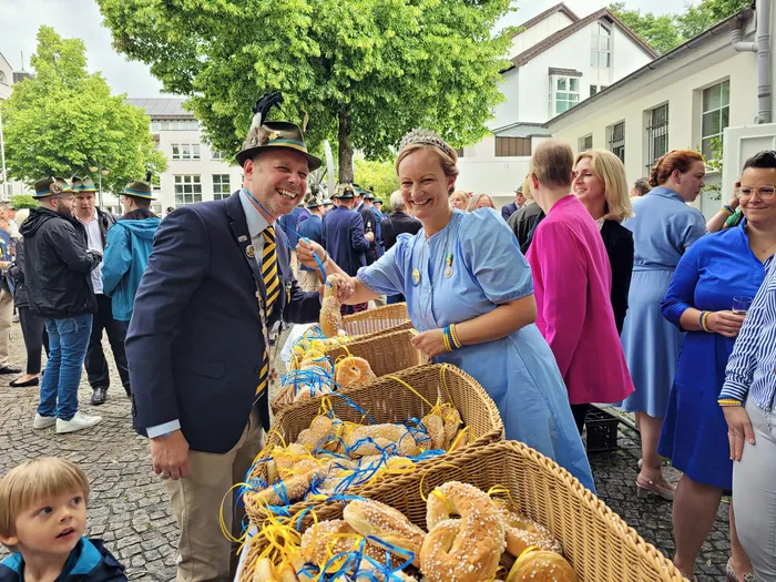 Ein Mann in Schützenuniform und eine Frau in hellblauem Kleid reichen sich bei einem Schützenfest die Hände. Vor ihnen stehen Körbe mit Brezeln.