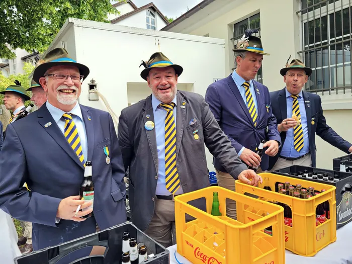 Vier Männer in blauen Anzügen und traditionellen Schützenhüten stehen an einem Tisch mit Getränkekisten. Einer von ihnen hält eine Zigarette, während ein anderer Flaschen aus einem Kasten nimmt.