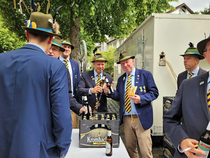 Mehrere Männer in traditionellen Schützenuniformen mit Hüten stehen um einen Tisch mit Bierkisten herum. Ein Mann hält zwei Flaschen Bier hoch, während andere ihm zuhören.